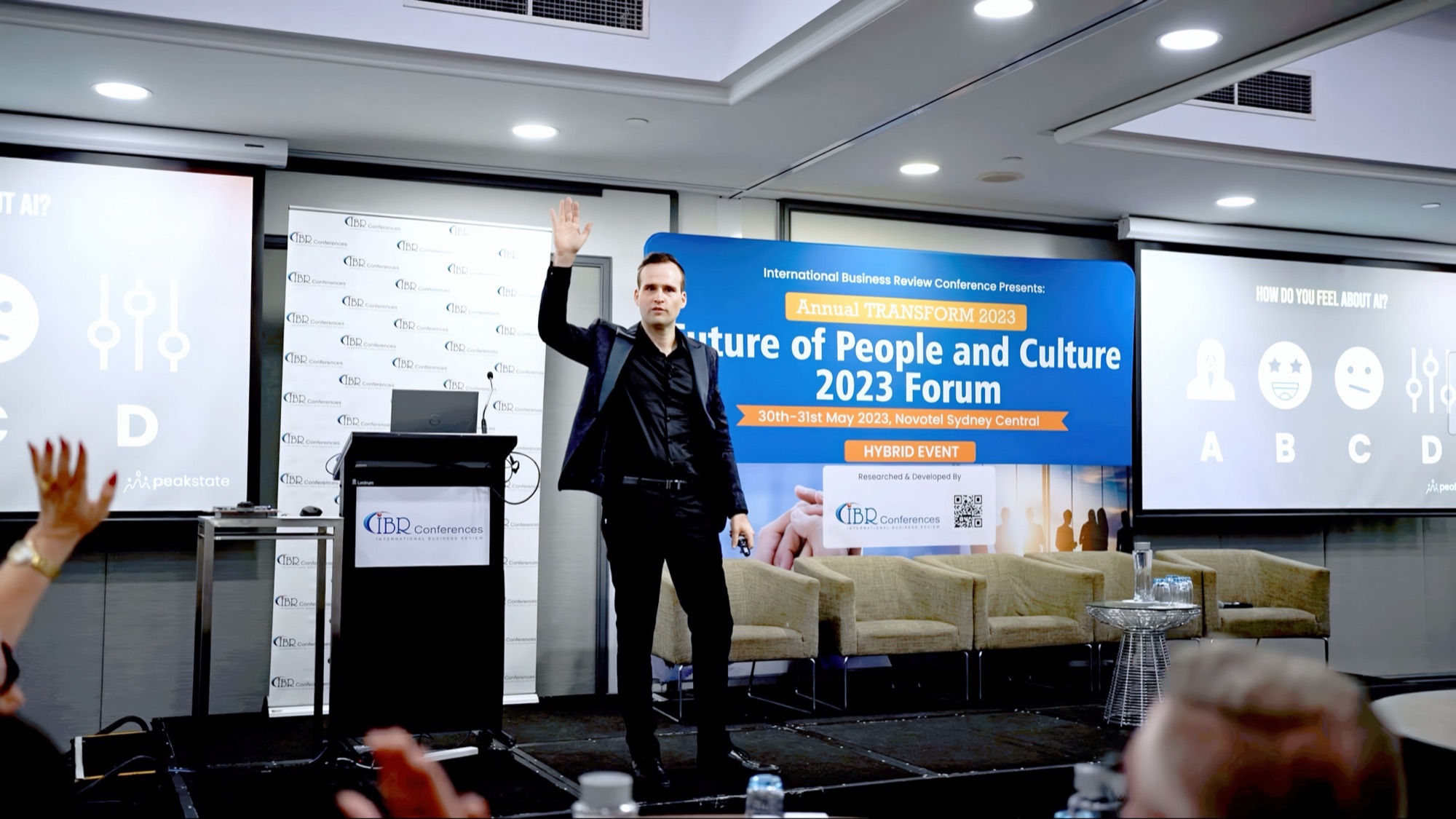 A speaker on stage at the Future of People and Culture 2023 Forum in Sydney, hand raised to the room.